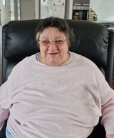 A woman in her 60's wearing a pale pink jumper and glasses, sitting in a dark leather armchair.