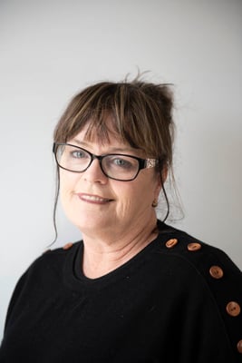 A woman wearing black-framed glasses and hair in a bun, wearing a black jumper with buttons on the shoulders.