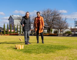 Two women in winter coats standing on a green lawn playing a skittle game.