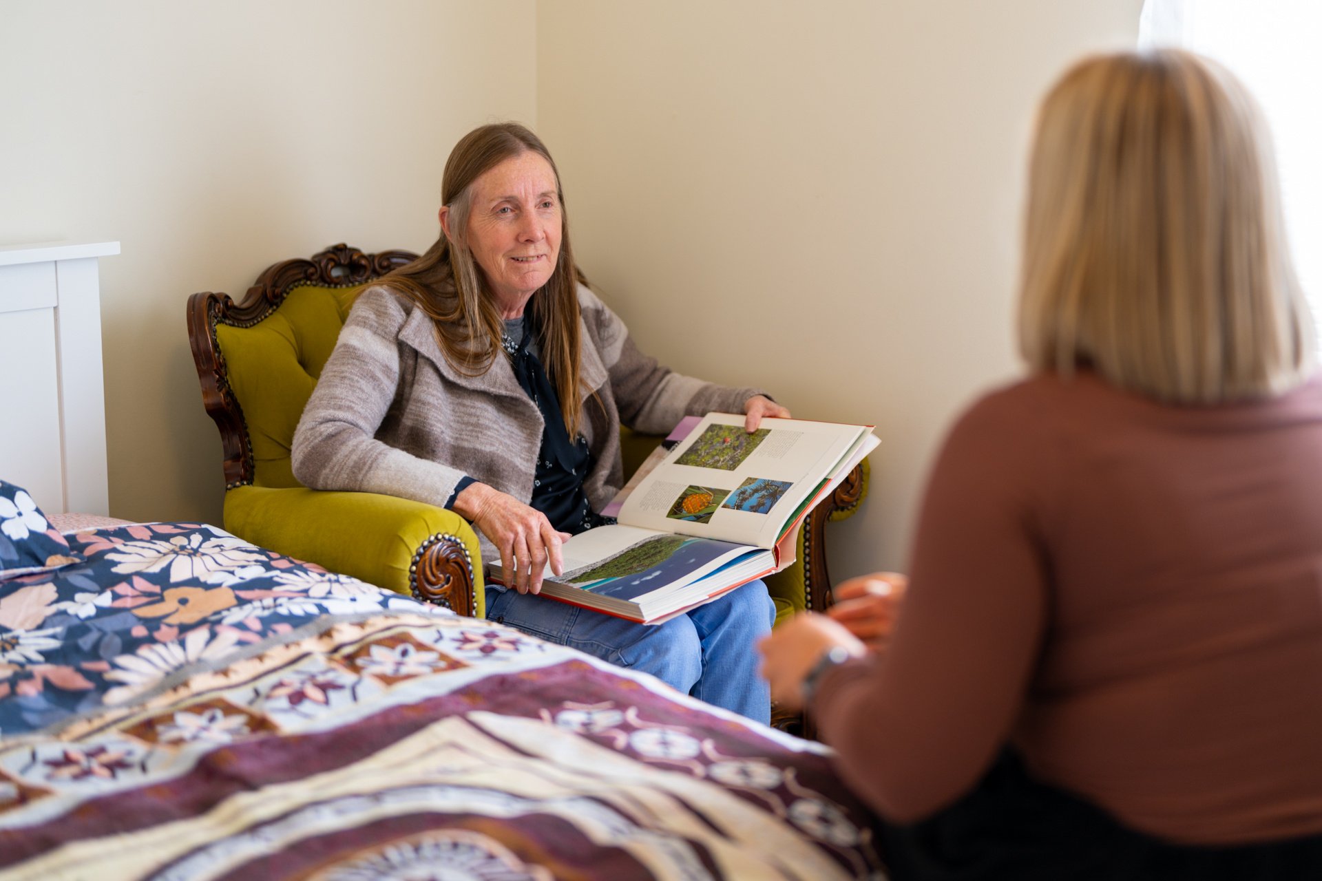 A woman in a green armchair holds an open book, engaged in conversation with someone seated nearby in a cozy bedroom with a floral bedspread.