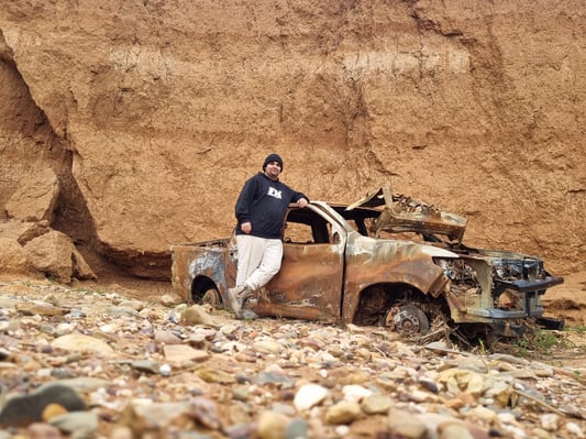 A young man in a black hoodie and beanie leaning against a rusted car wreck.