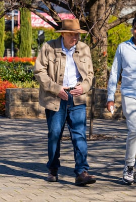 A man in a jacket and jeans wearing a broad brimmed hat, walking along a path.