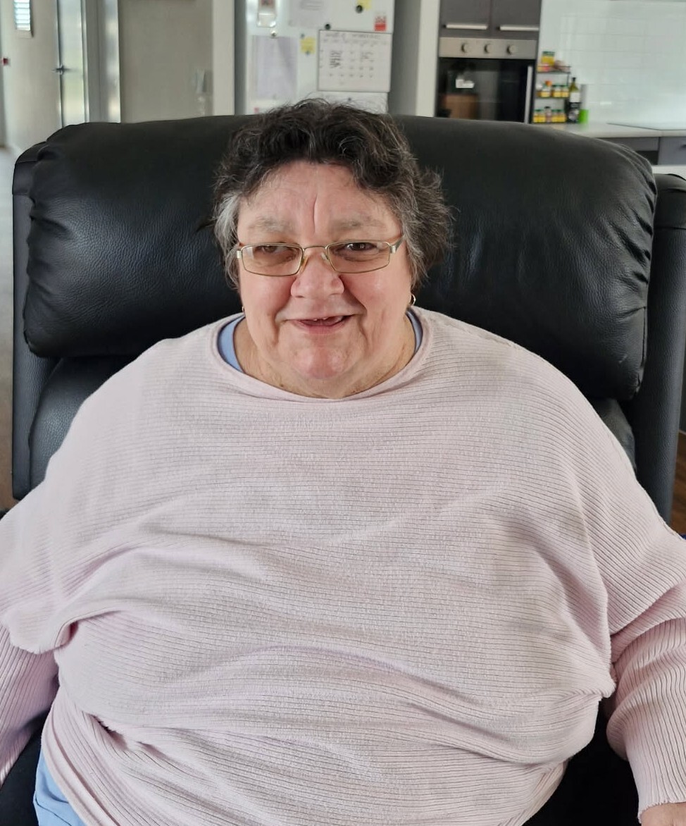 A woman in her 60's wearing a pale pink jumper and glasses, sitting in a dark leather armchair.