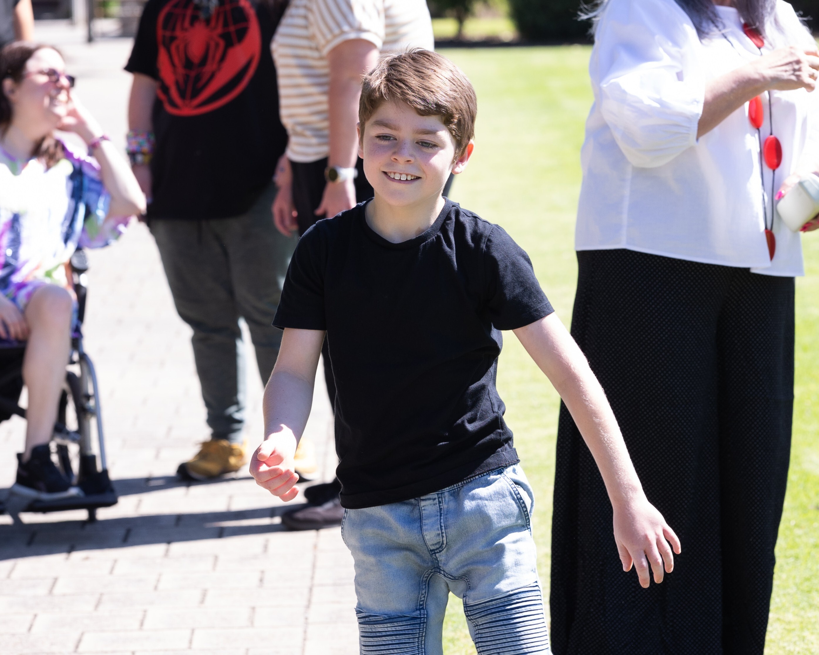 ID: photo of boy in black shirt and denim shorts smiling outside on a sunny day