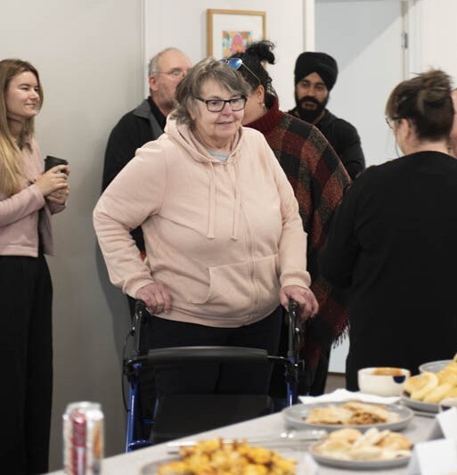 A woman in her 60's in a light pink jumper and glasses moving through a group of people while using a walker.
