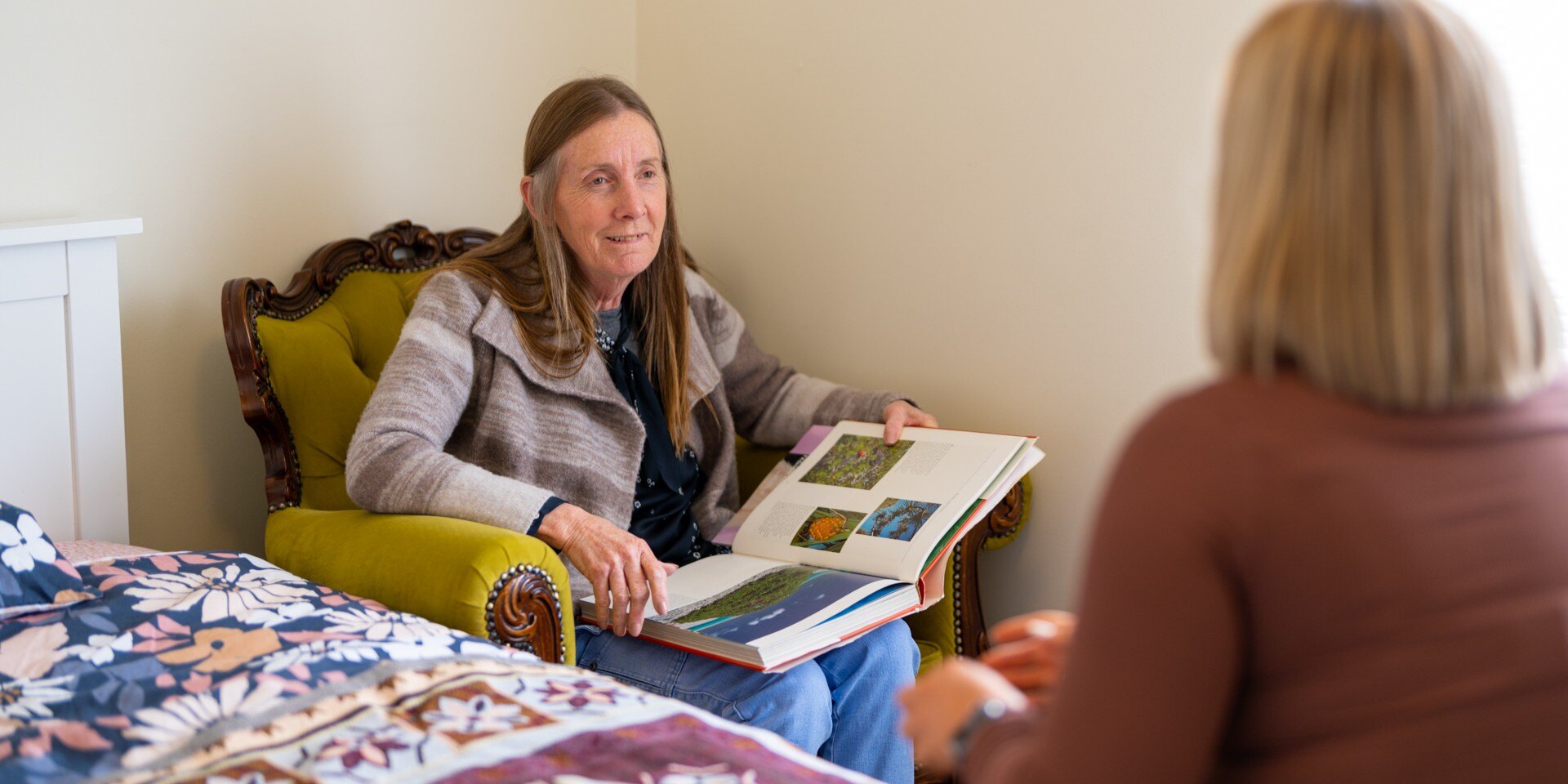 A woman on a green armchair holds an open book, engaged in conversation with someone seated nearby in a cozy bedroom with a floral bedspread.