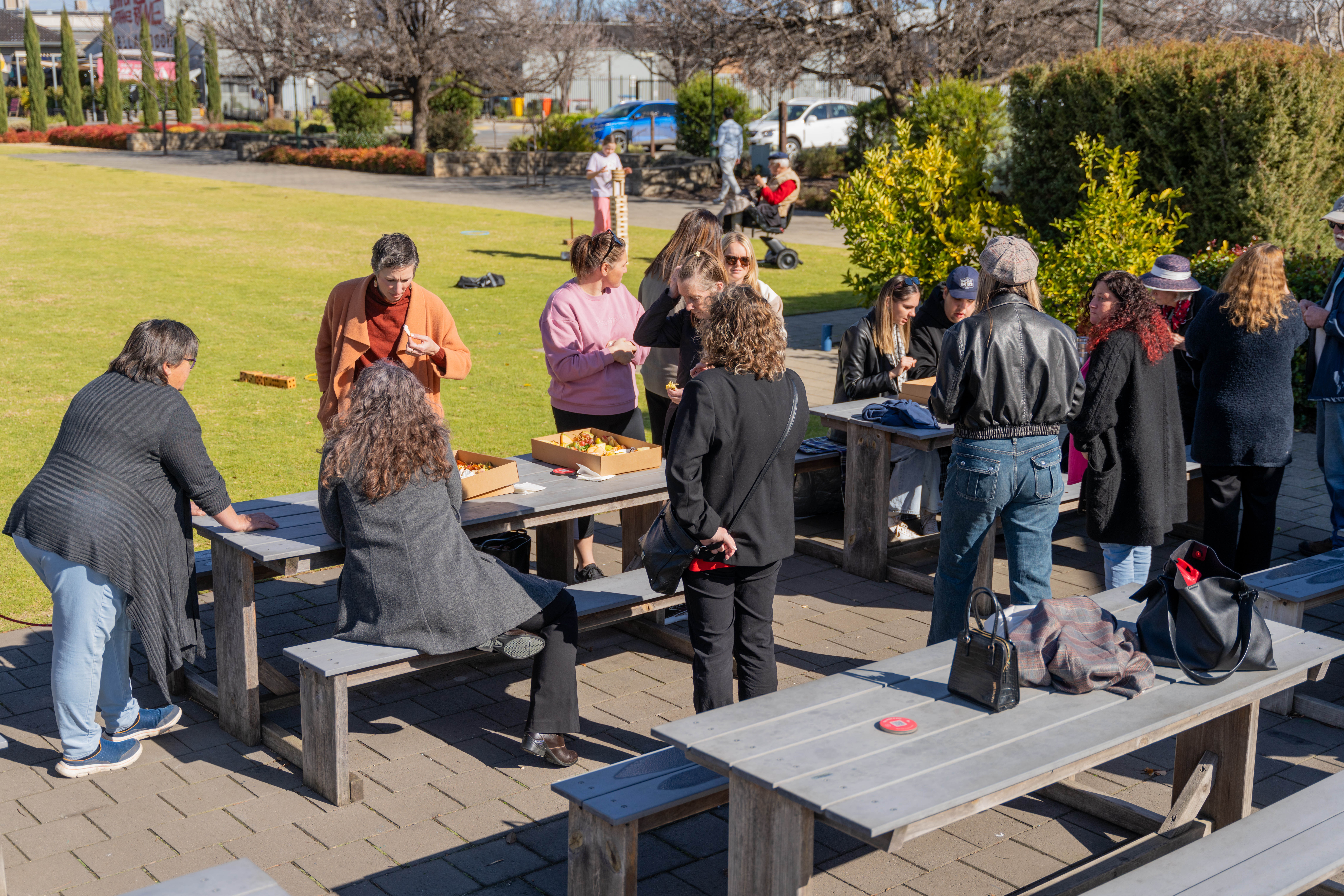 A large gathering of people standing amongst picnic tables beside a big lawned area.