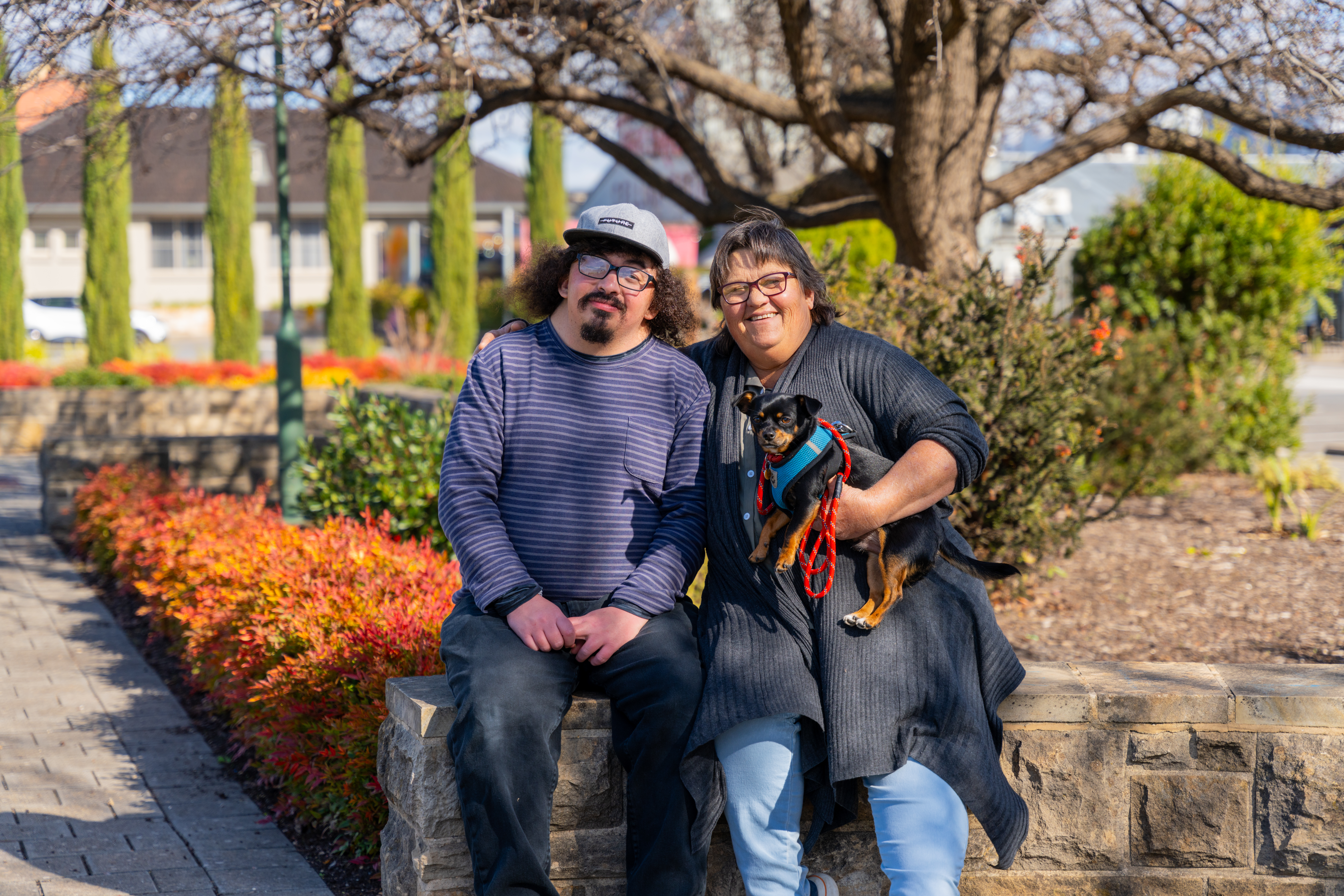 **Alt Text:** A cheerful outdoor scene featuring two people sitting on a low stone wall surrounded by vibrant landscaping with red and green foliage. On the left, a man in a striped long-sleeve shirt and glasses smiles warmly. On the right, a woman in a dark cardigan and glasses holds a small black-and-brown dog wearing a red harness. 