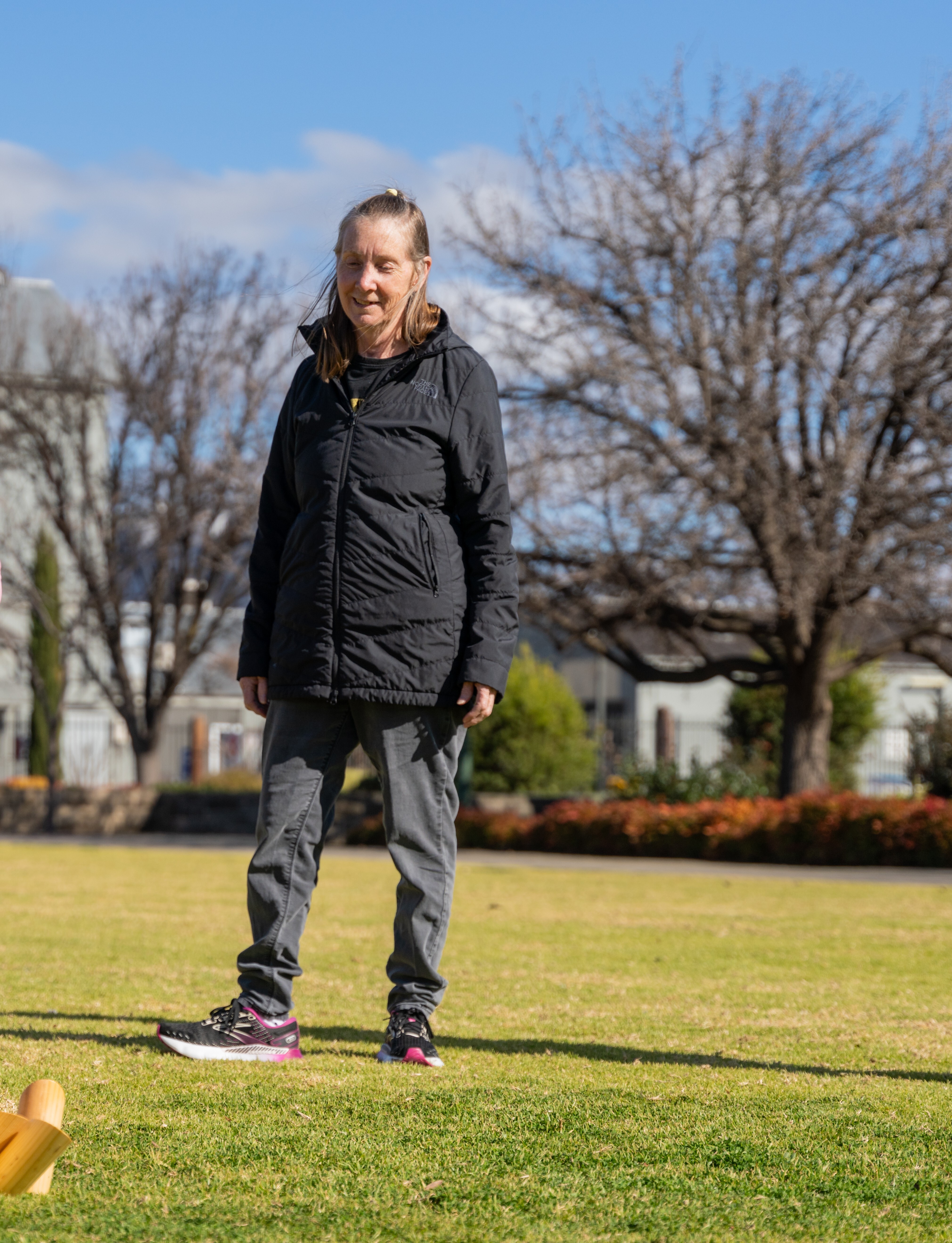 A blonde woman in her 60's wearing a black jacket standing on a green lawn with bare winter trees in the background.